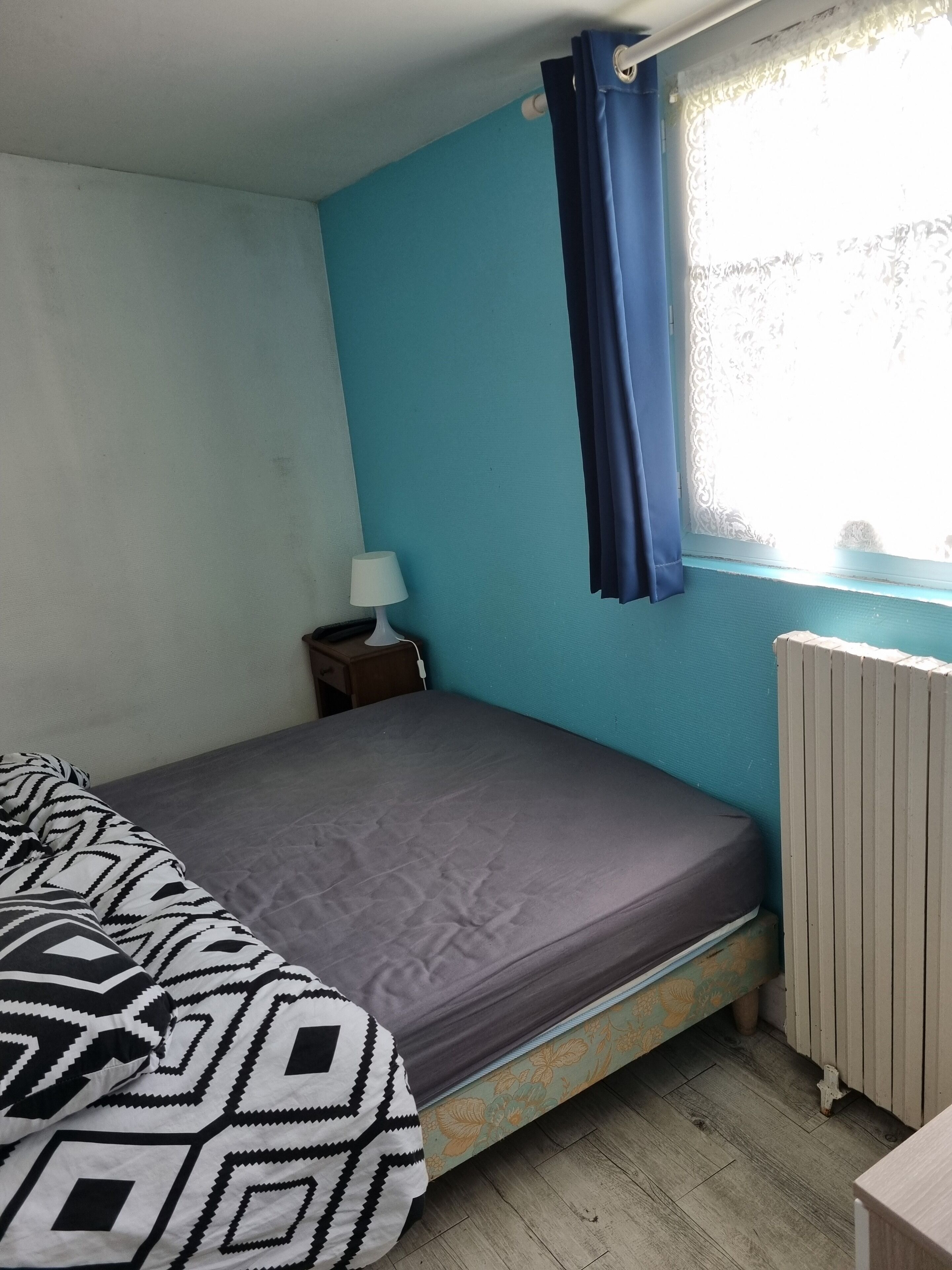 Una stanza piccola con un letto singolo, parete blu, finestra con tenda, comodino con lampada e termosifone.