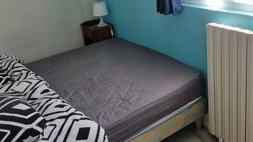Una stanza piccola con un letto singolo, parete blu, finestra con tenda, comodino con lampada e termosifone.