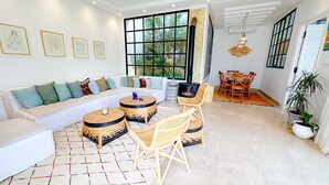 A modern living room with a white sofa, wicker side tables, and a dining area with a table and chairs. - Villa Azur Piscine Bord de mer a Hammamet (Hammamet)