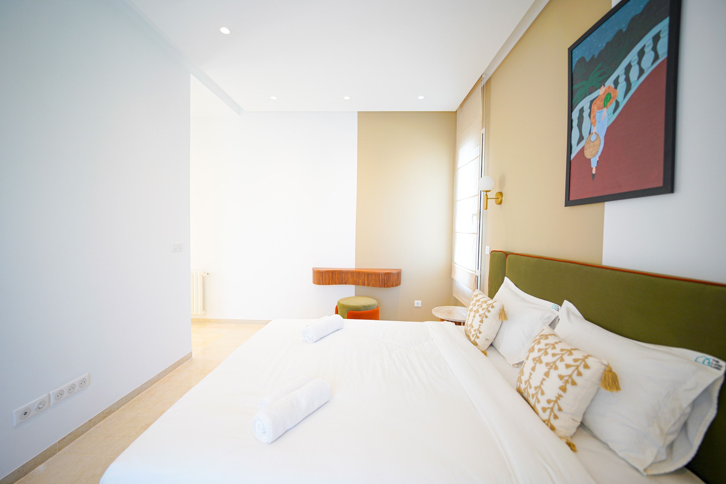 A hotel room with a large bed, a small stool, a nightstand, and a painting on the wall.