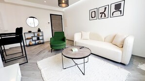 Ein modernes Wohnzimmer mit einem grünen Sessel, einem weißen Sofa, einem runden Couchtisch und einem schwarzen Stuhl. An der Wand hängt ein Spiegel, eine Regalfläche zeigt dekorative Gegenstände und im Hintergrund ist eine Tür zu sehen.