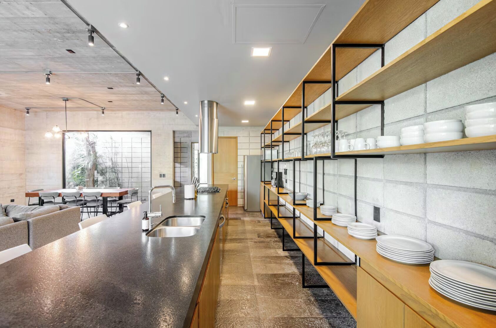A modern kitchen with a long counter, built-in shelves with dishes, and a view of the outdoors.