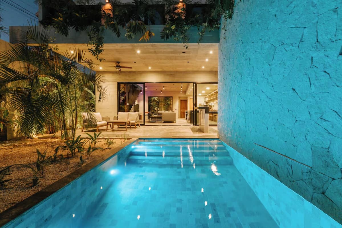 A modern outdoor pool area with a wooden deck, lounge chairs, and a view into the house.