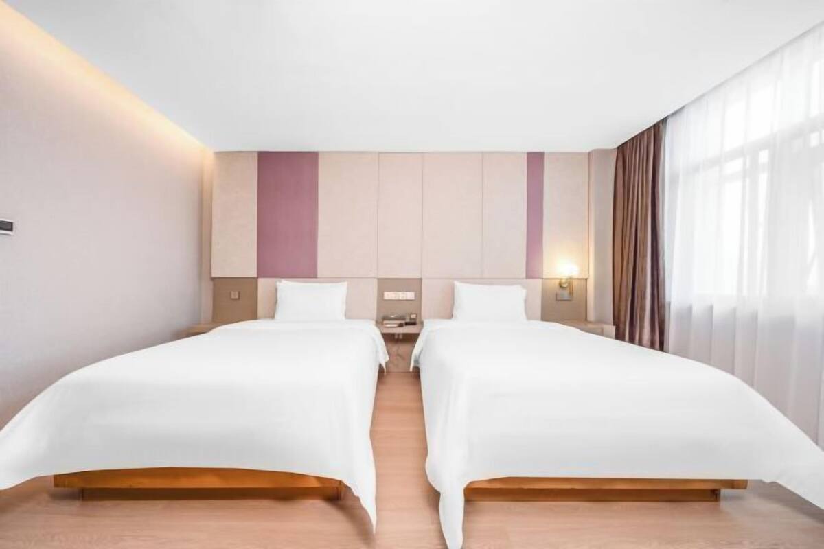 A hotel room with two beds, a wooden floor, a large window with curtains, and a wall-mounted light.