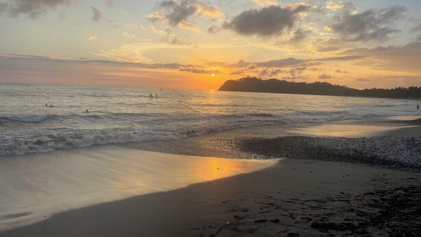 Une plage avec des vagues, un coucher de soleil et une terre lointaine à l’horizon.