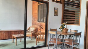 Une pièce dotée d’une porte-fenêtre coulissante donnant sur un balcon, d’un banc en bois, d’une chaise en osier, d’une table à manger avec des chaises, et d’une fenêtre avec des stores en bois.