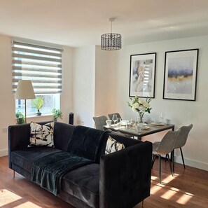 A modern living room with a dark sofa, a dining table set for two, and a pendant light. - The Hidden Story - Manchester (Greater Manchester)