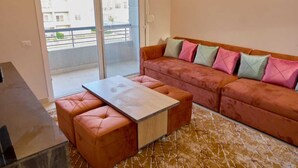 A modern living room with a sofa and ottoman, a glass door leading to a balcony, and a patterned rug. - Long Island Chalets Family only (Marsa Matrouh)