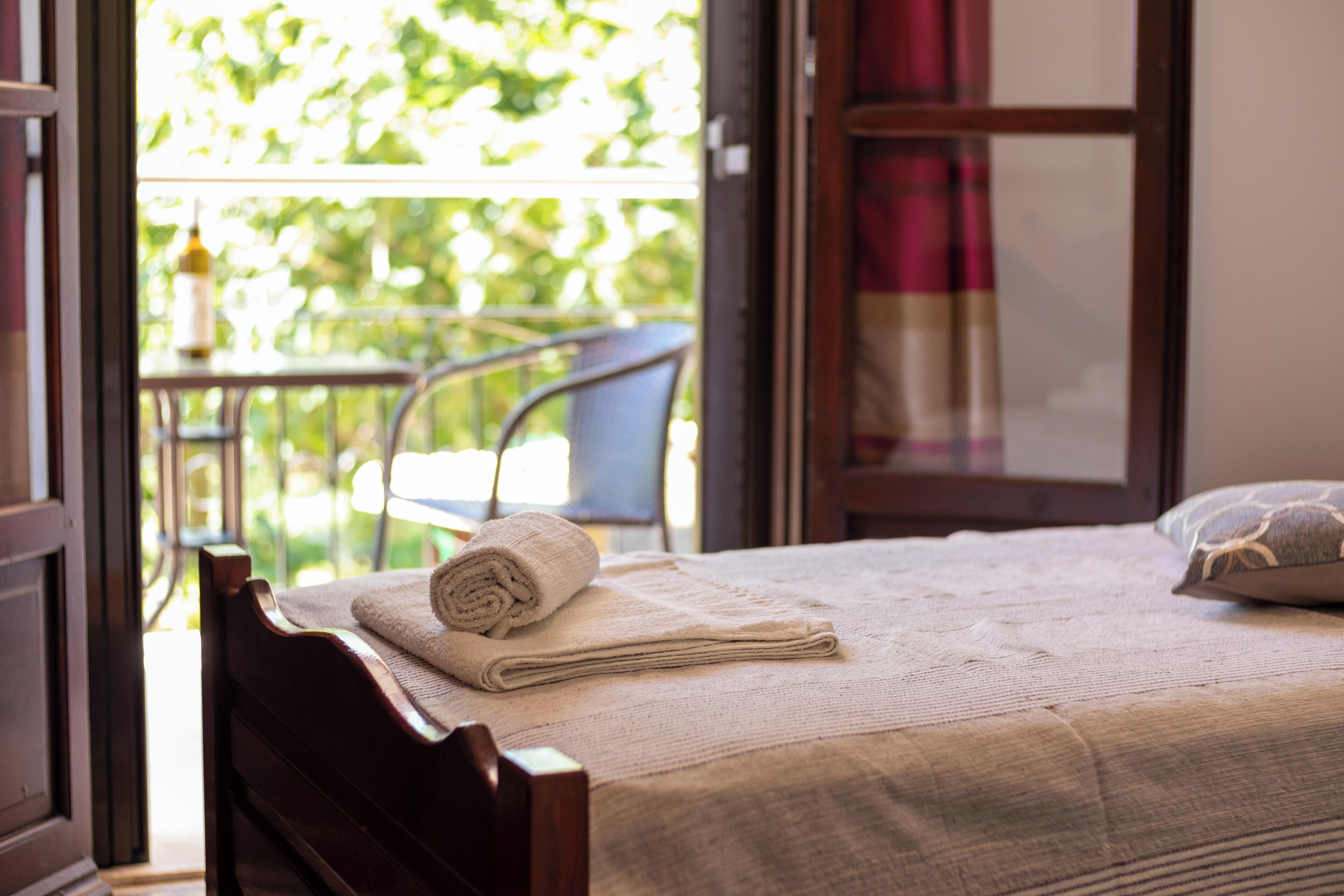 A bed with a rolled towel and a pillow, a wooden headboard, and a view of greenery through a window.