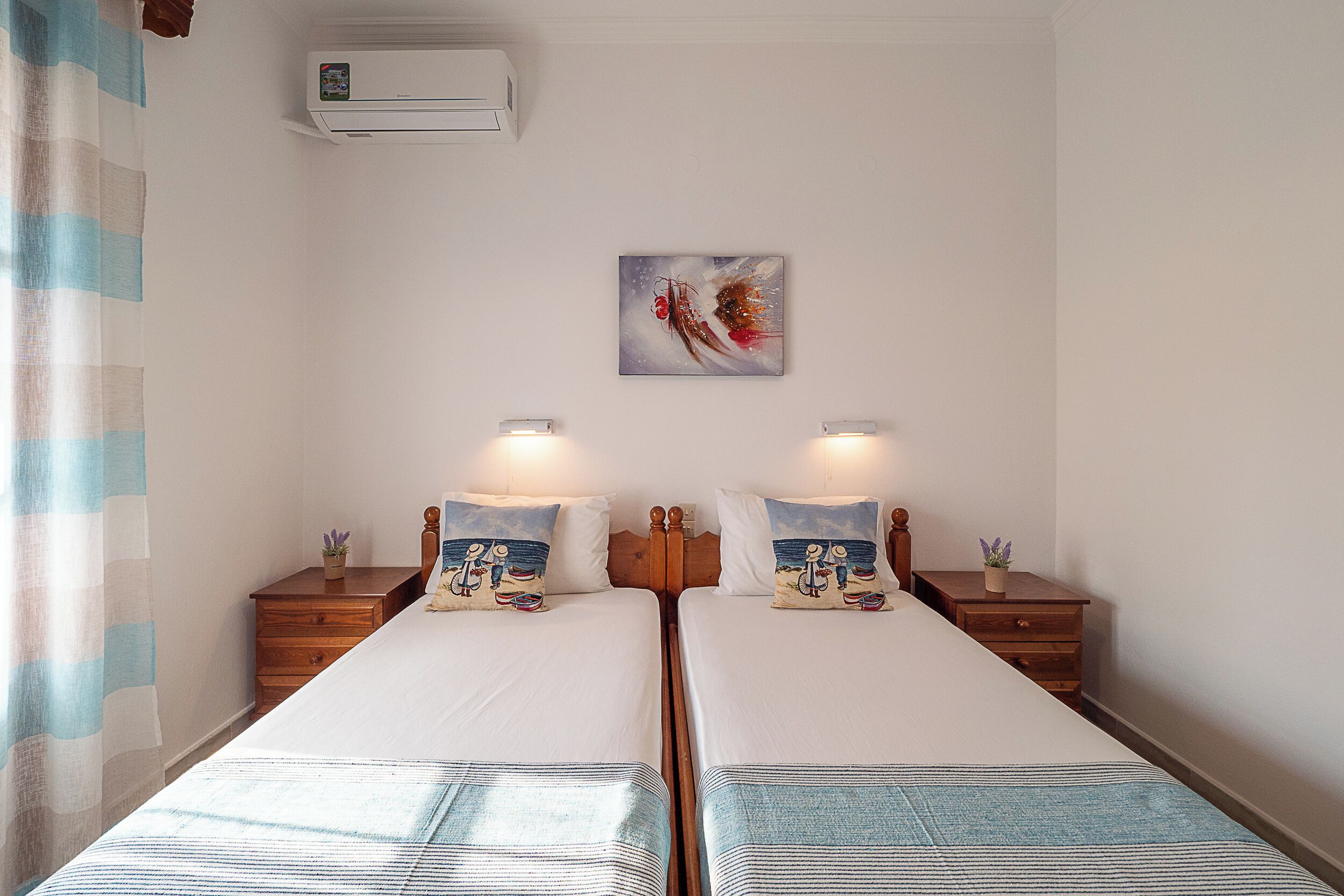 Une chambre avec deux lits, chacun doté d’une tête de lit et d’une table de chevet assorties. Un climatiseur mural est fixé au mur, et un tableau est accroché au-dessus des lits.