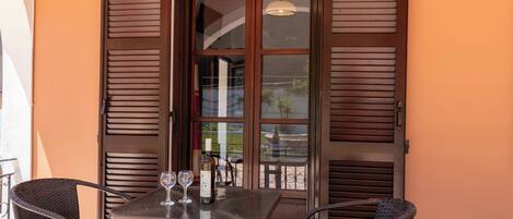 Una mesa con dos sillas, una botella de vino, una ventana con contraventanas de madera y una lámpara colgante.