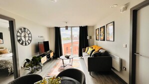 Ein modernes Wohnzimmer mit einer Couch, einem Couchtisch und einem Fernseher. Es gibt ein großes Fenster mit Vorhängen, eine Uhr an der Wand und eine Heizung.