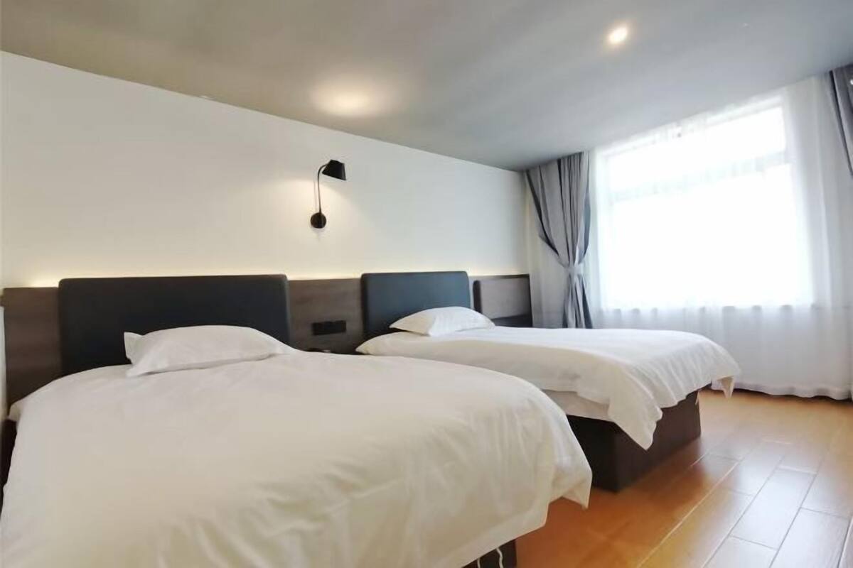 Habitación de hotel con dos camas, una mesita de noche, una lámpara de pared y un ventanal con cortinas.