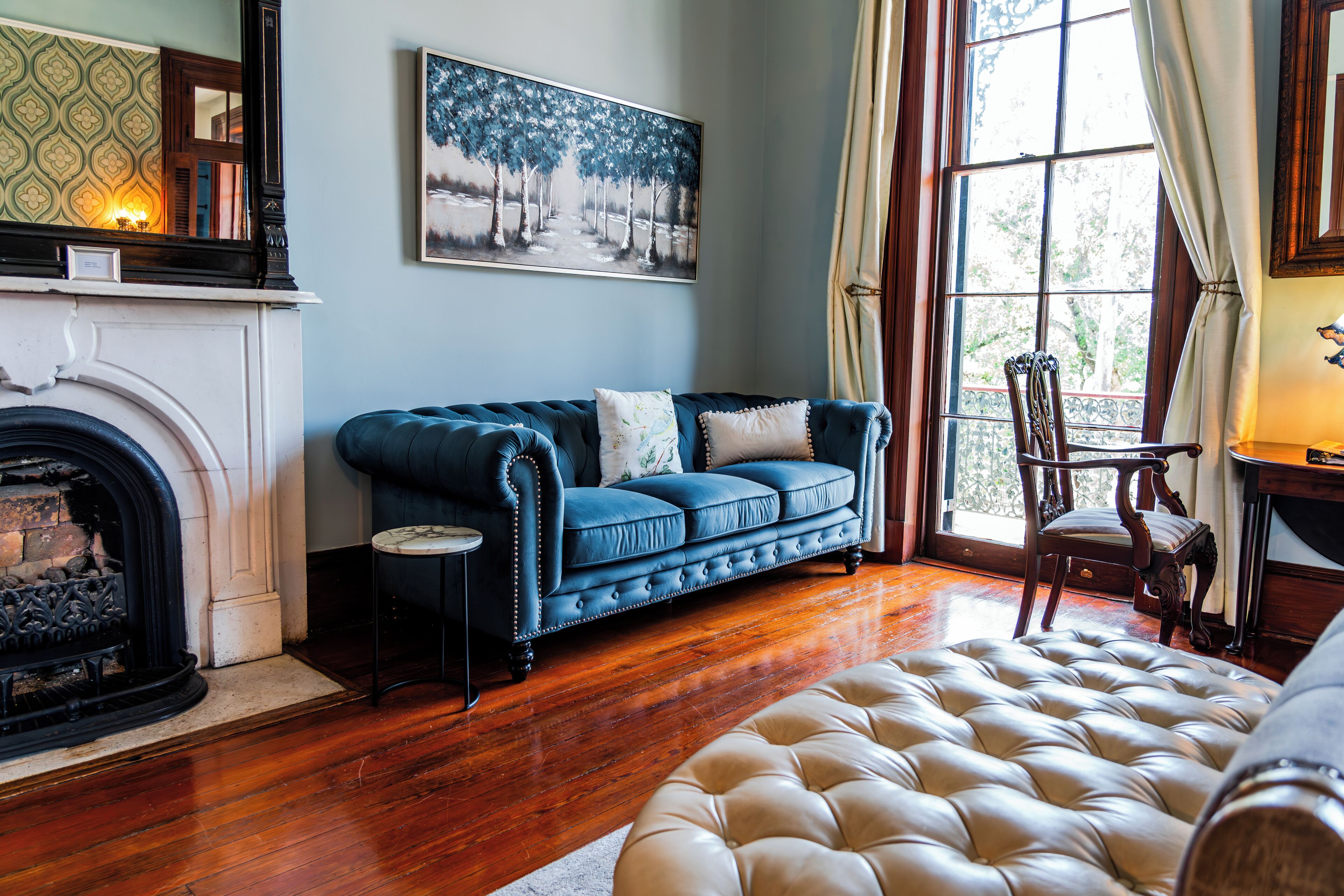 Une pièce avec une cheminée, un canapé bleu, un grand pouf moelleux et un tableau représentant des arbres.
