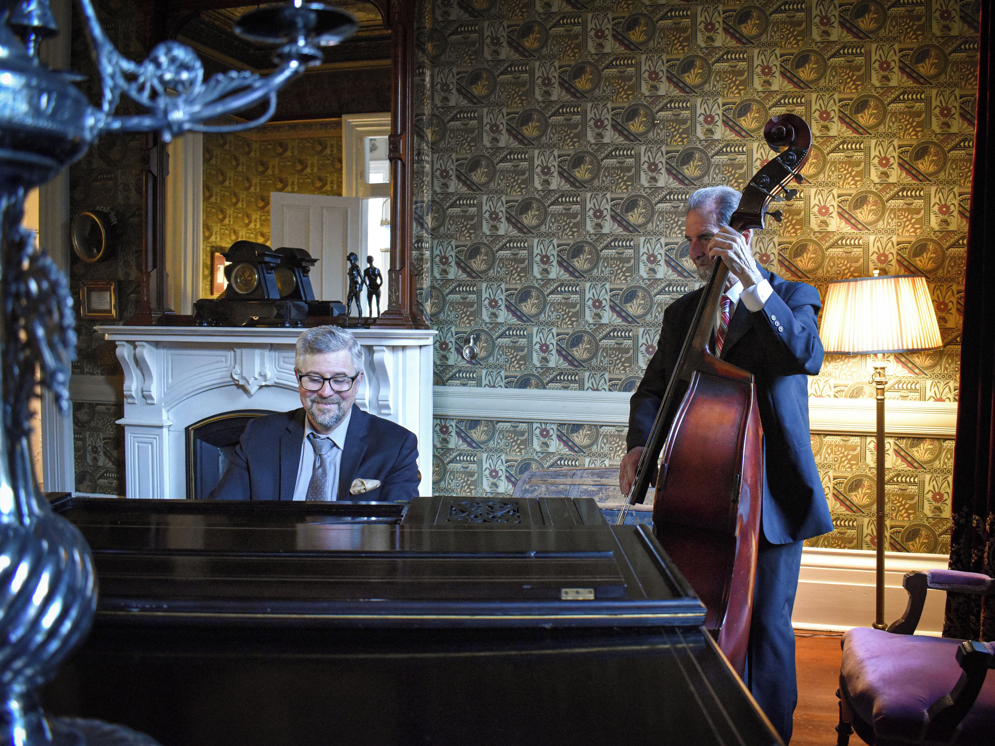 Un homme joue du violoncelle dans une pièce où se trouvent un piano, une cheminée et un papier peint à motifs.