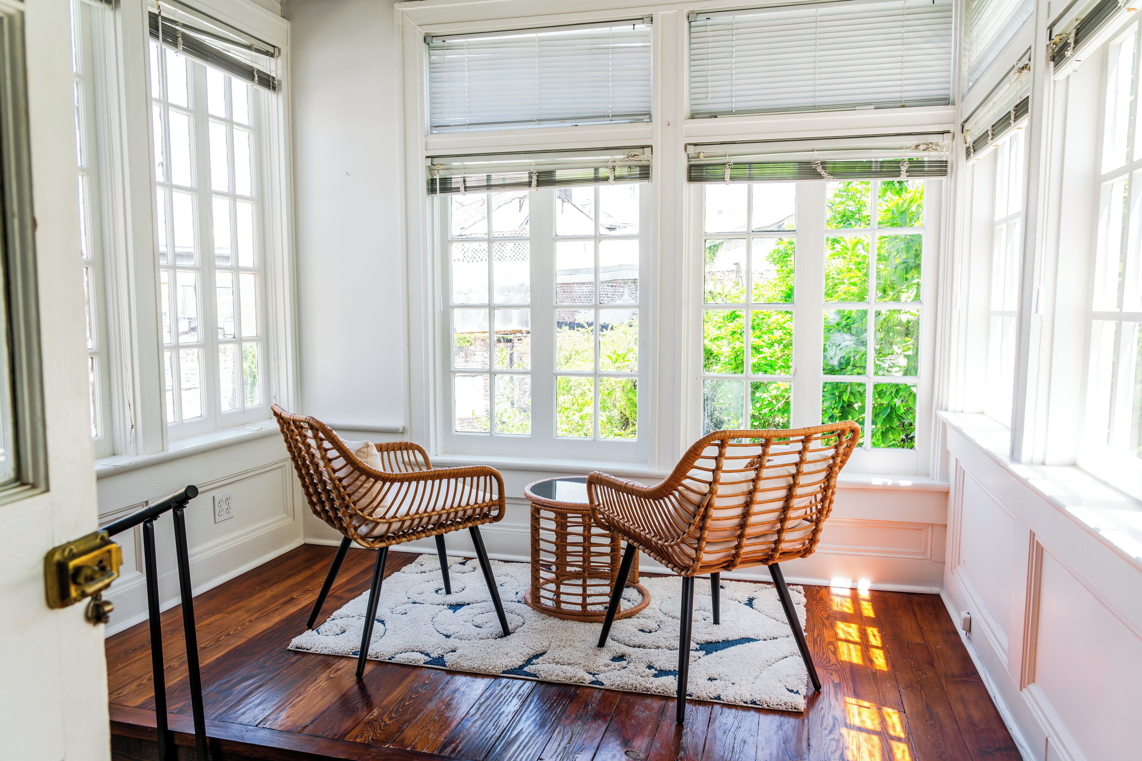Una habitación con piso de madera, dos sillas de mimbre, una mesita redonda y ventanas grandes con persianas.