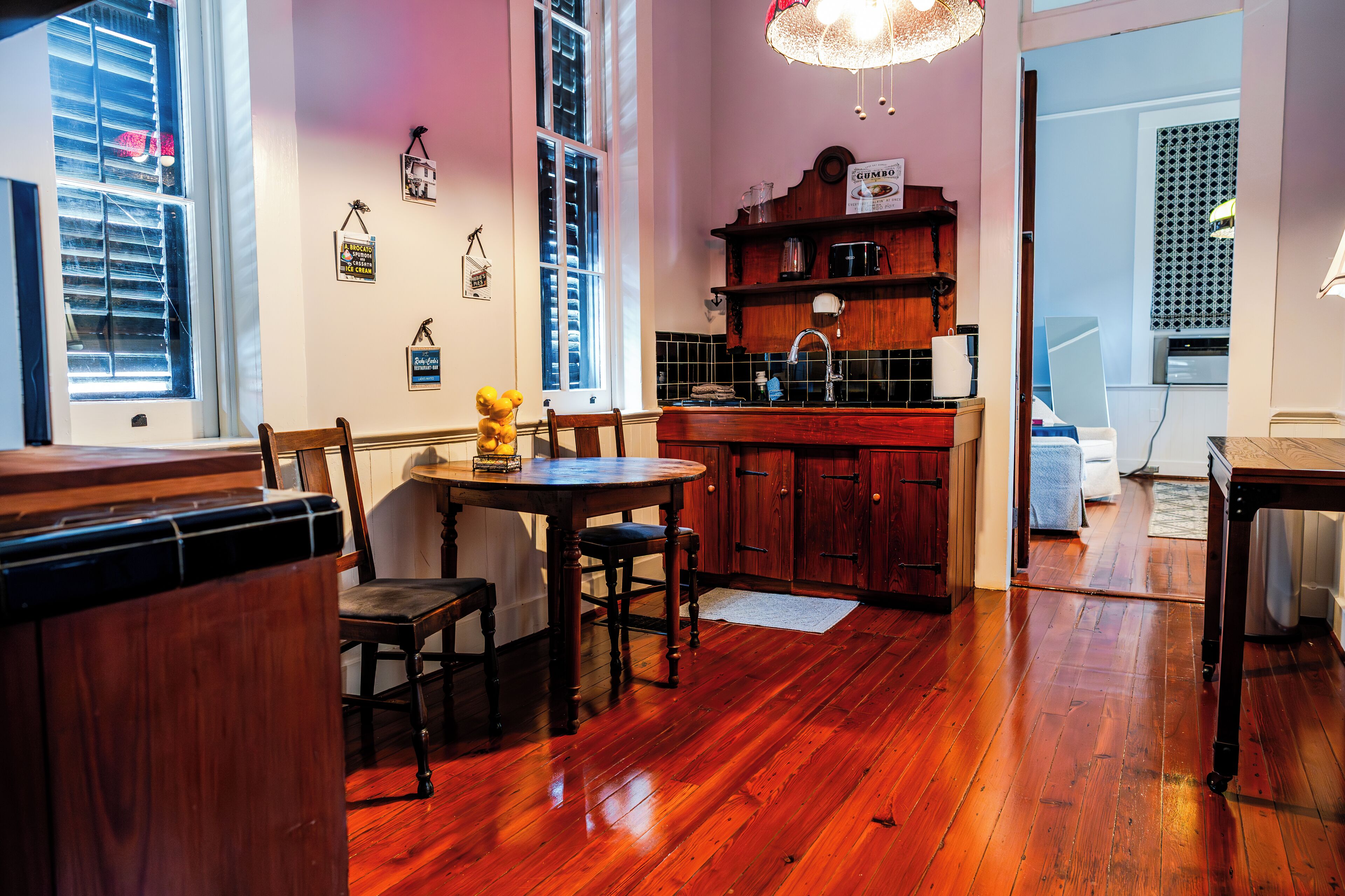 A cozy kitchen with wooden cabinets, a dining table, and chairs. There is a window with blinds, a hanging light fixture, and a small shelf with kitchen items.
