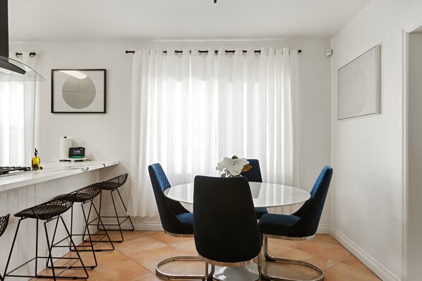 Uma área de jantar moderna com uma mesa redonda, quatro banquetas altas, um vaso branco com uma flor e uma cortina branca.