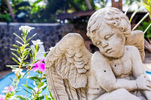 Una estatua de ángel de piedra con tallas intrincadas, ubicada cerca de una piscina rodeada de vegetación.