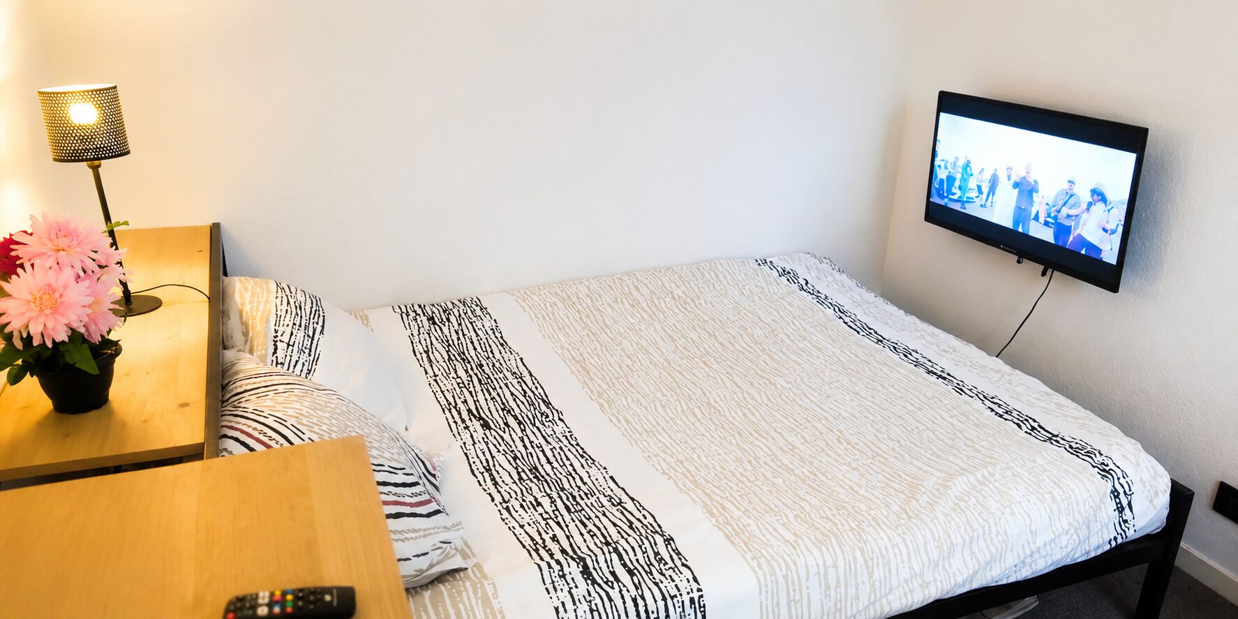 Uma cama de solteiro com colcha estampada, uma mesa de cabeceira com luminária e flores, e uma televisão presa na parede.