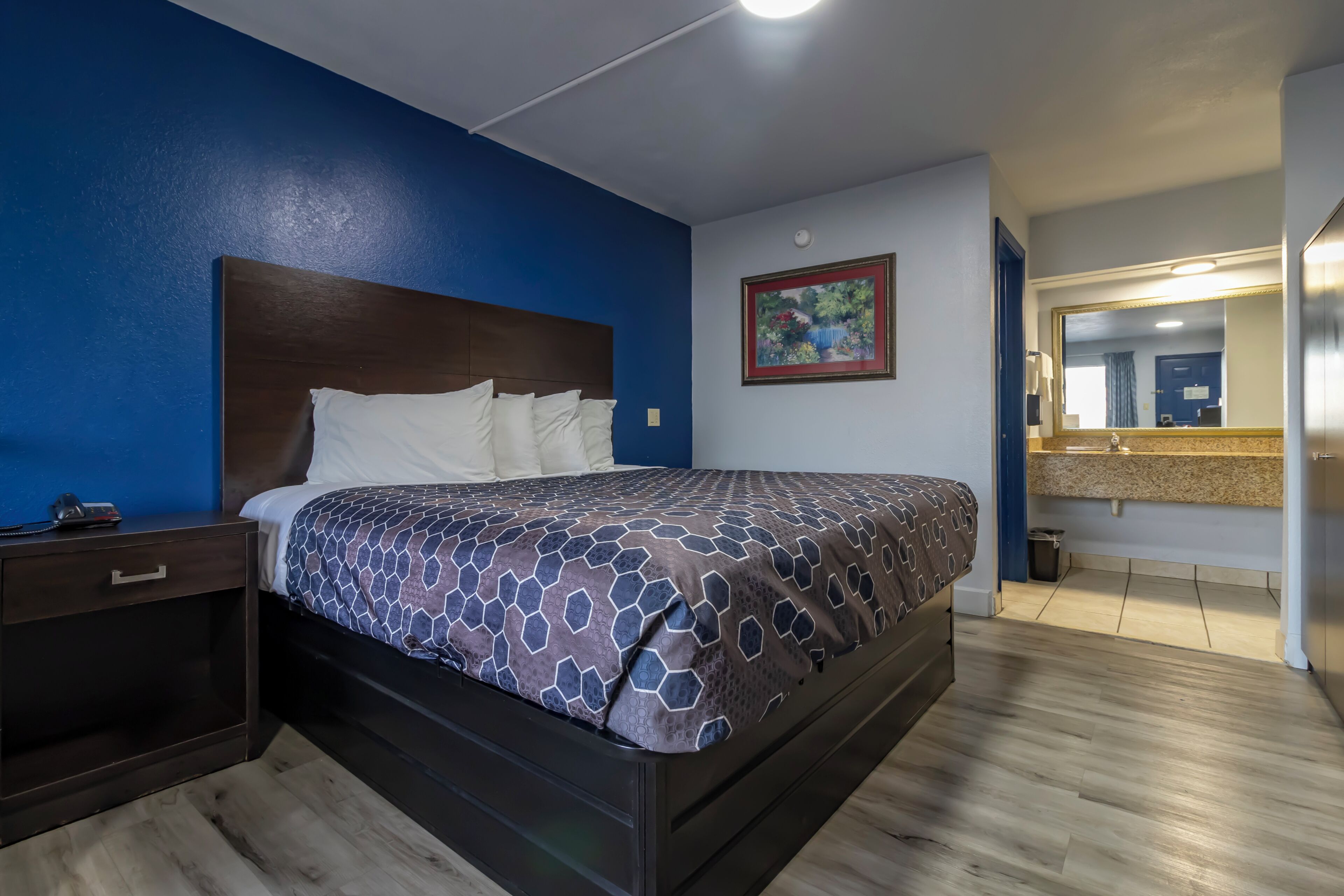 Une chambre d’hôtel avec un lit, une table de chevet, un tableau, un miroir et une salle de bain visible à travers une porte ouverte.
