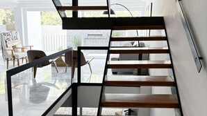 Una escalera moderna con barandillas de vidrio y peldaños de madera, que conduce a un área de estar con un sofá y sillones.