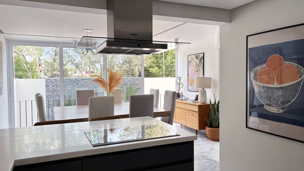 Una cocina moderna con una isla grande, comedor con sillas, un armario de madera y vistas al exterior.