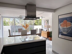 A modern kitchen with a large island, dining area with chairs, a wooden cabinet, and a view of greenery outside.