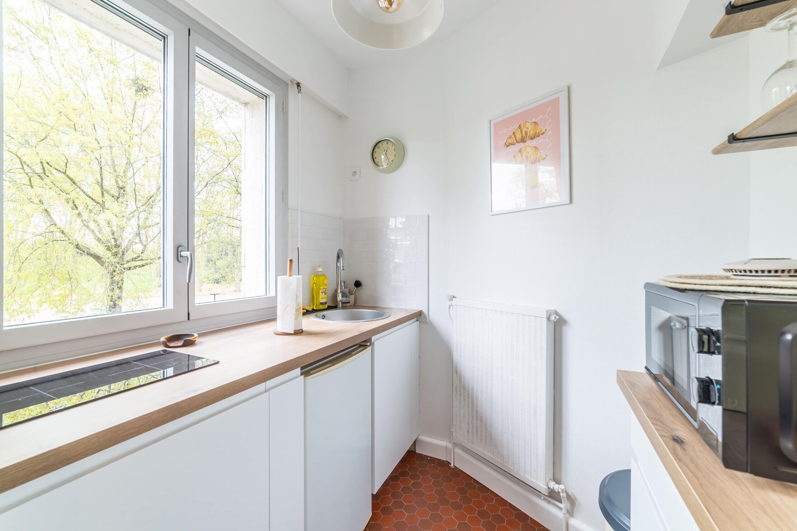 A kitchen with a window, white cabinets, a sink, a clock, and a radiator.
