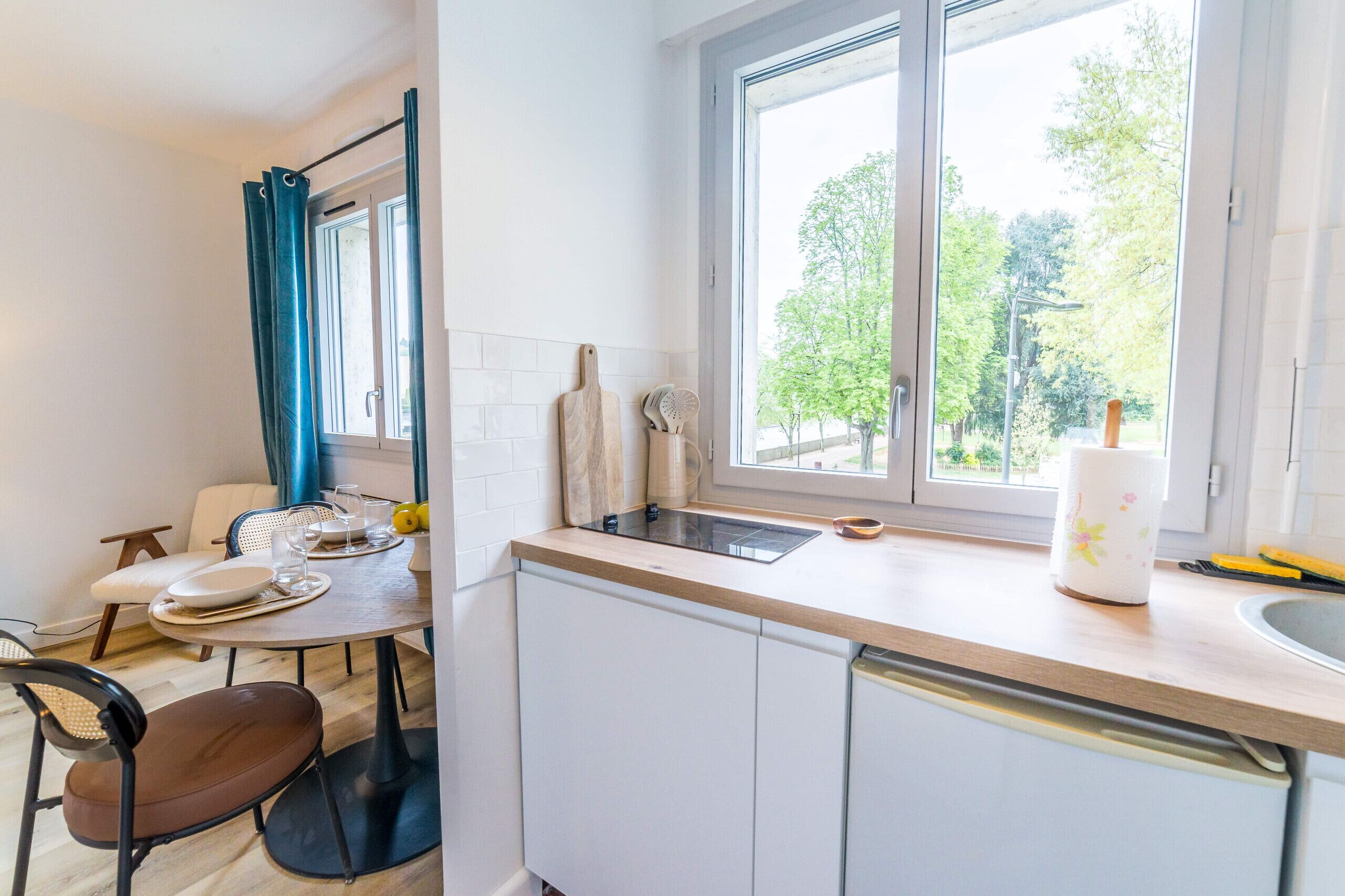 A kitchen with white cabinets, a window with a view of trees, a dining table with chairs, and a countertop with a sink and utensils.