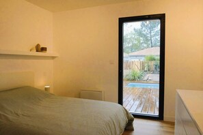 A bedroom with a bed, a bedside table, a radiator, and a view of an outdoor deck and pool. - Modern house with a pool, nestled between the lake and the forest (Lacanau)
