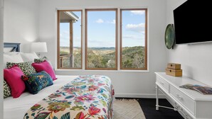 A bedroom with a bed, a desk, and a large window with a view of the countryside.