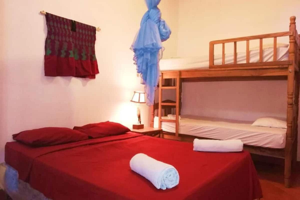 A room with two beds, one with a red bedspread and the other with a white one. There is a rolled white towel on the bed with the red bedspread.
