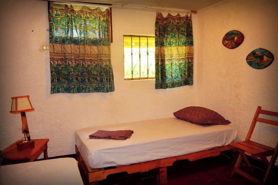 Una habitación pequeña y sencilla con una cama individual, una ventana con rejas y dos cuadros colgados en la pared.