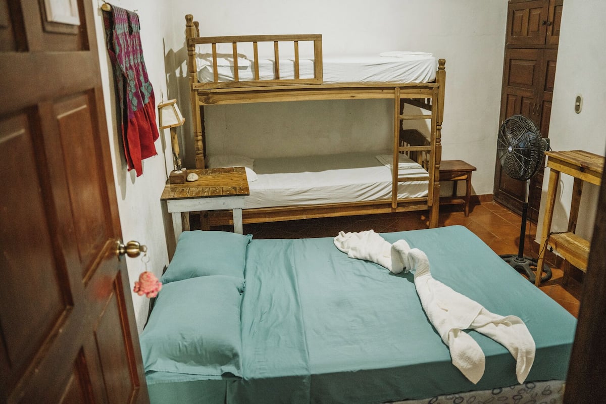 A dormitory room with two bunk beds, a wooden table, a fan, and a door.