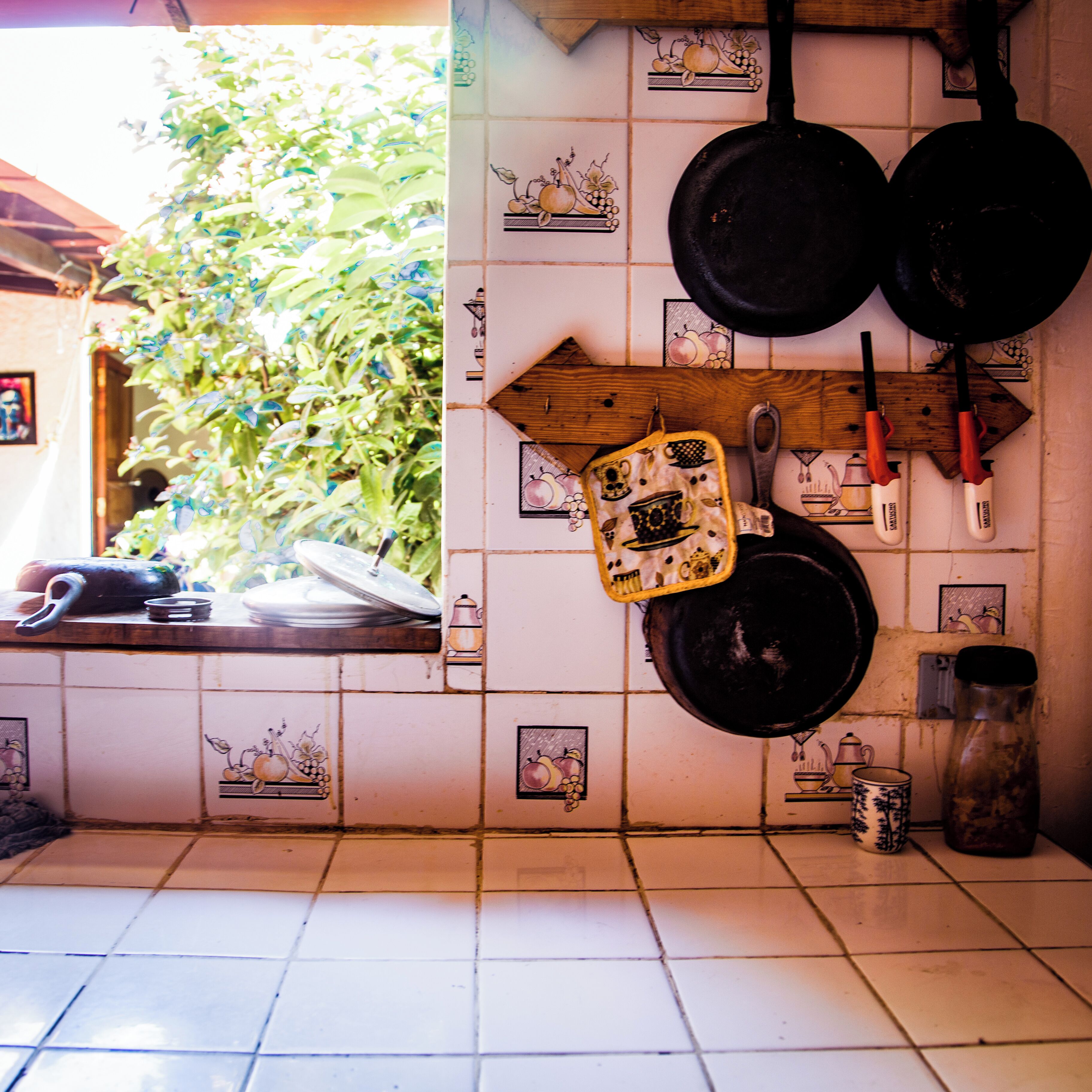 A kitchen with white tiles featuring fruit illustrations, hanging pots, and a wooden shelf with kitchen utensils.
