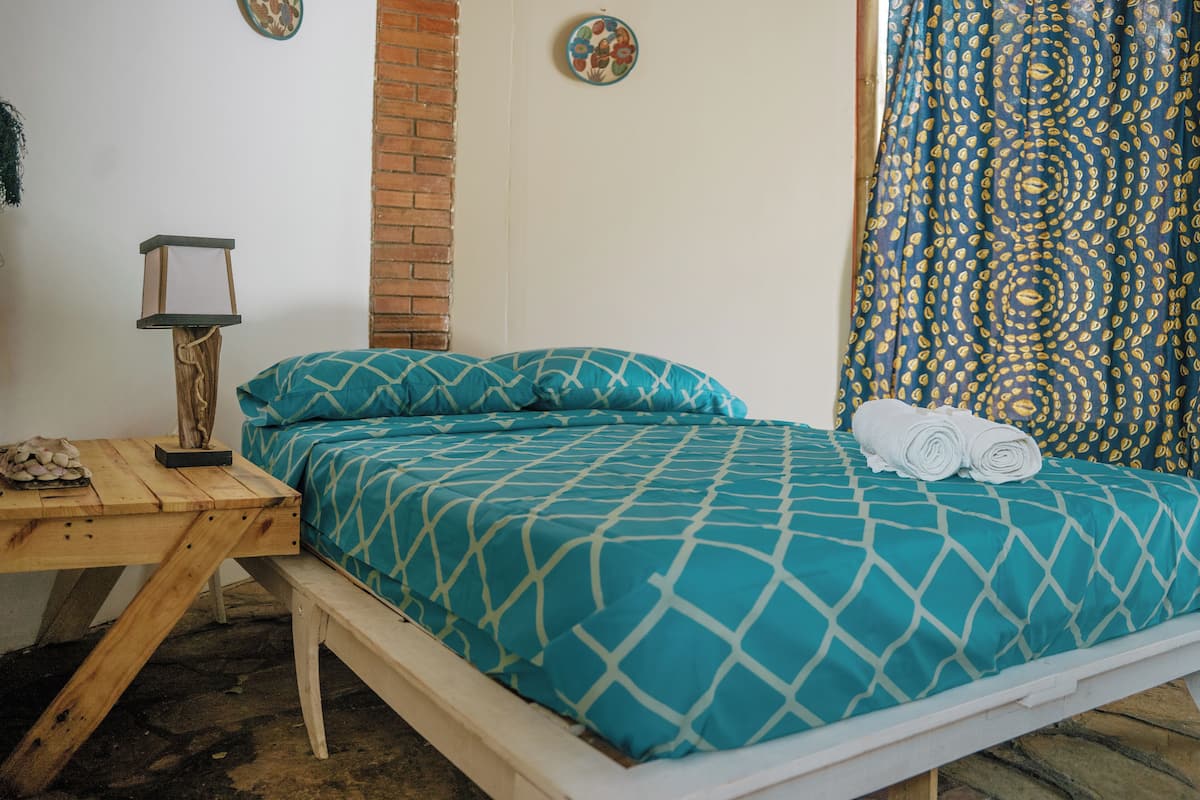 A bed with teal patterned bedding, a wooden nightstand with a lamp, and a brick wall.