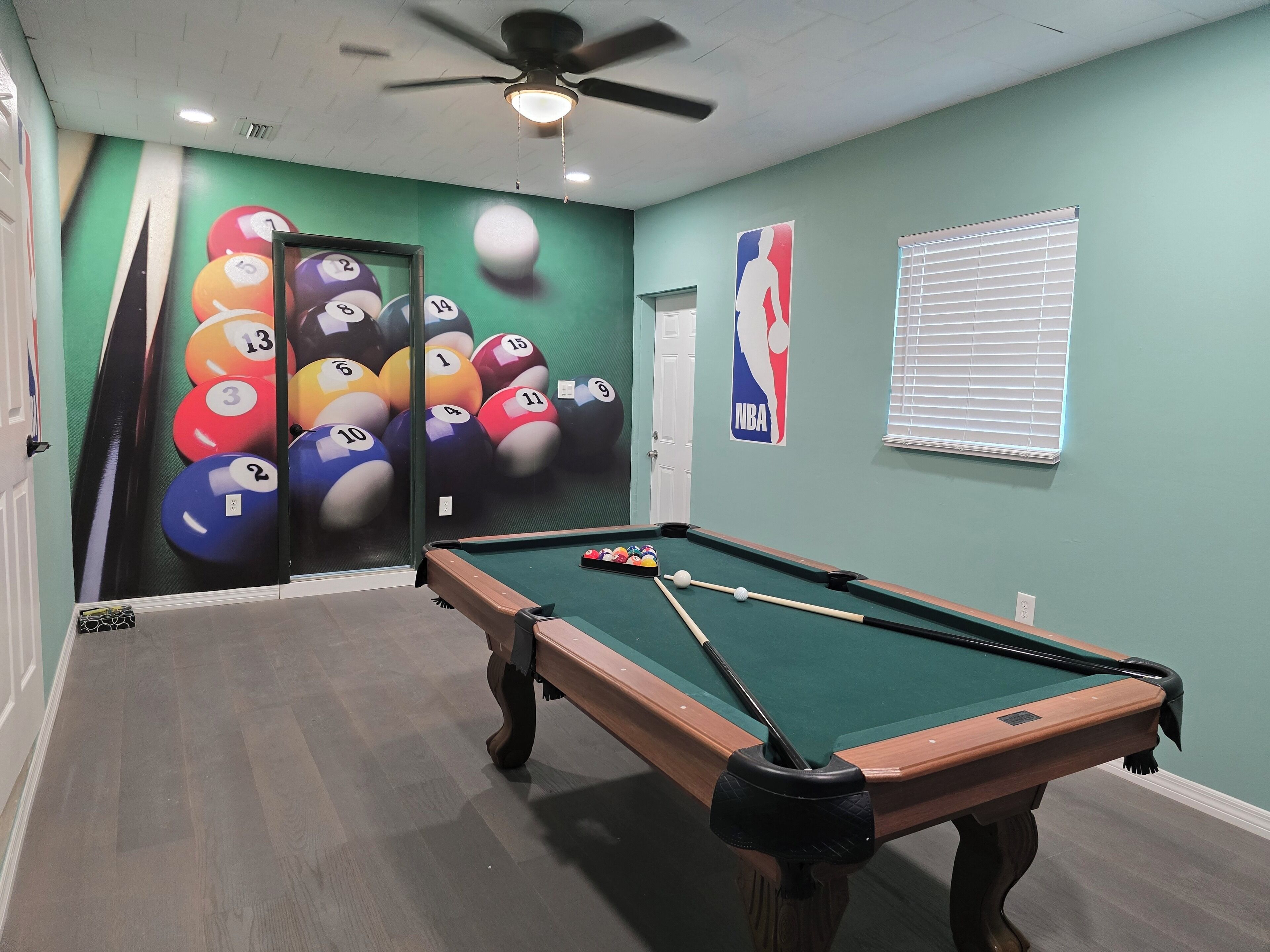 Een pooltafel in een ruimte met groene wanden, een plafondventilator en een NBA-logo.