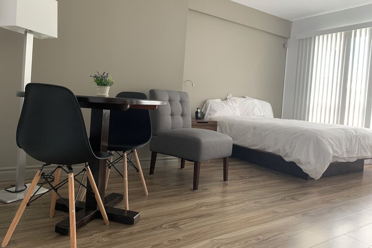 Ein modernes Hotelzimmer mit Holzboden, einem Bett mit weißer Bettwäsche, einem grauen Sessel, einem schwarzen Bürostuhl, einem kleinen Tisch mit Topfpflanze und einem Nachttisch mit Lampe.