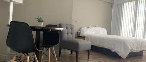 A modern hotel room with a wooden floor, a bed with white bedding, a grey armchair, a black office chair, a small table with a potted plant, and a bedside table with a lamp.
