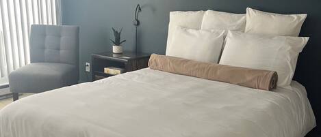 A bedroom with a large bed, a chair, a nightstand with a lamp, and a wall-mounted picture.
