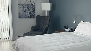 A modern bedroom with a large bed, a chair, a bedside table with a lamp, and a painting on the wall. - HĂ´tel Normandie (Rimouski)