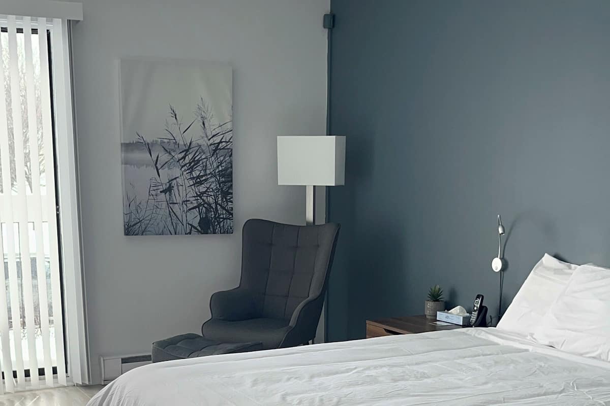 Ein modernes Schlafzimmer mit einem großen Bett, einem Sessel, einem Nachttisch mit Lampe und einem Bild an der Wand.