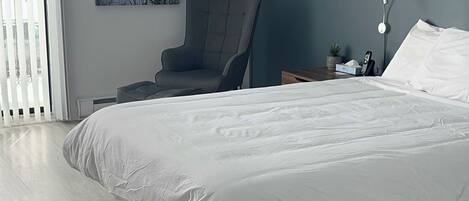 A modern bedroom with a large bed, a chair, a bedside table with a lamp, and a painting on the wall.