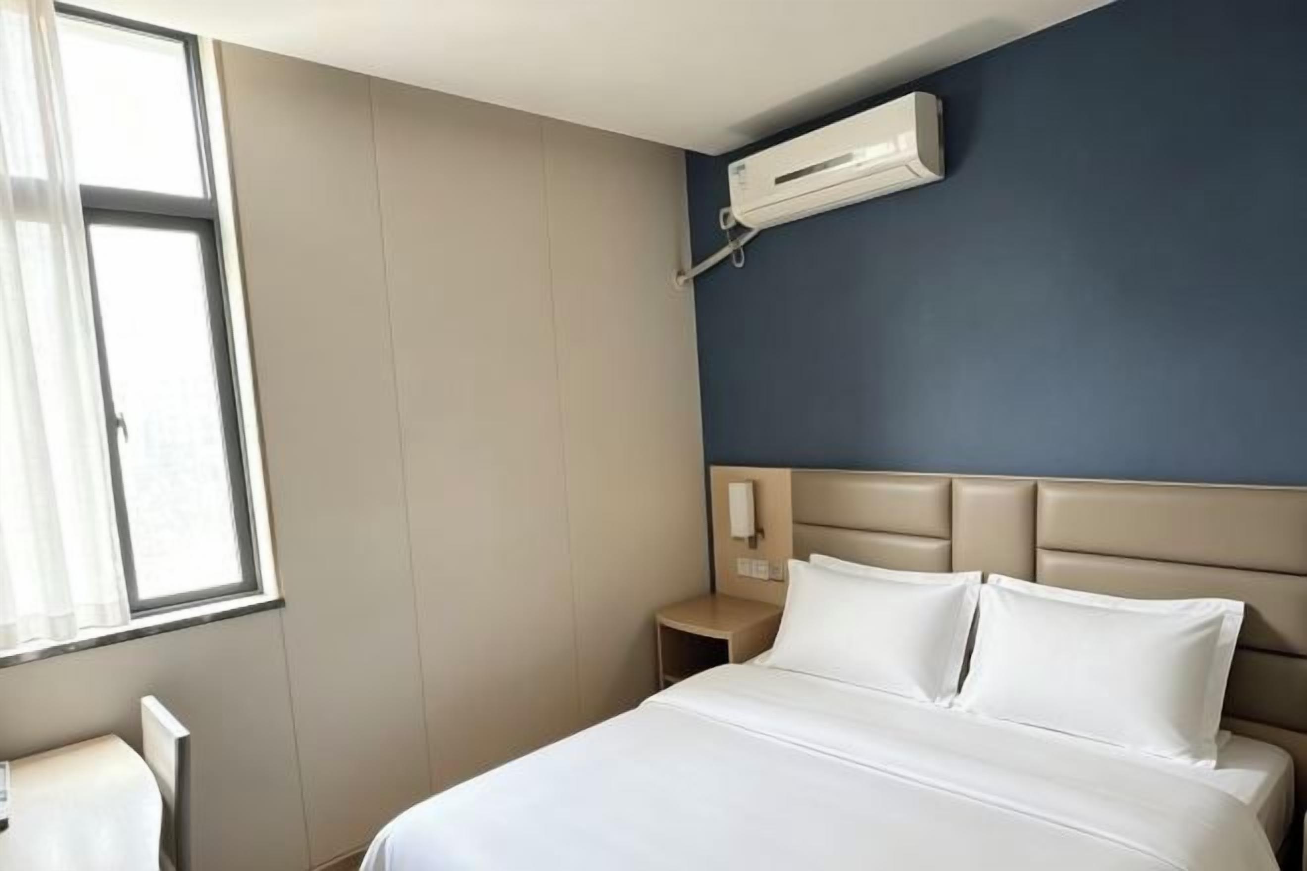 A hotel room with a bed, a nightstand, a wall-mounted air conditioner, and a window.