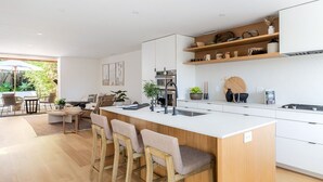 A modern kitchen with a central island, bar stools, a dining area with a table and chairs, and a view of greenery outside.
