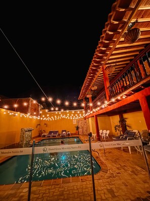 Un espace piscine d’hôtel avec une terrasse en bois, des chaises longues et des guirlandes lumineuses.