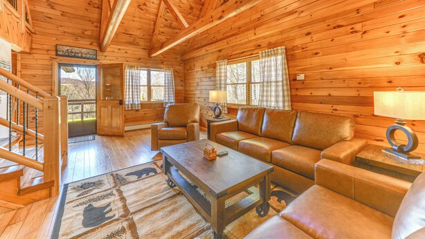 A wooden cabin interior with a staircase, leather sofa, and a coffee table. - Modern Comforts Classic Cabin: Family/Fish Escape! (Deposit)