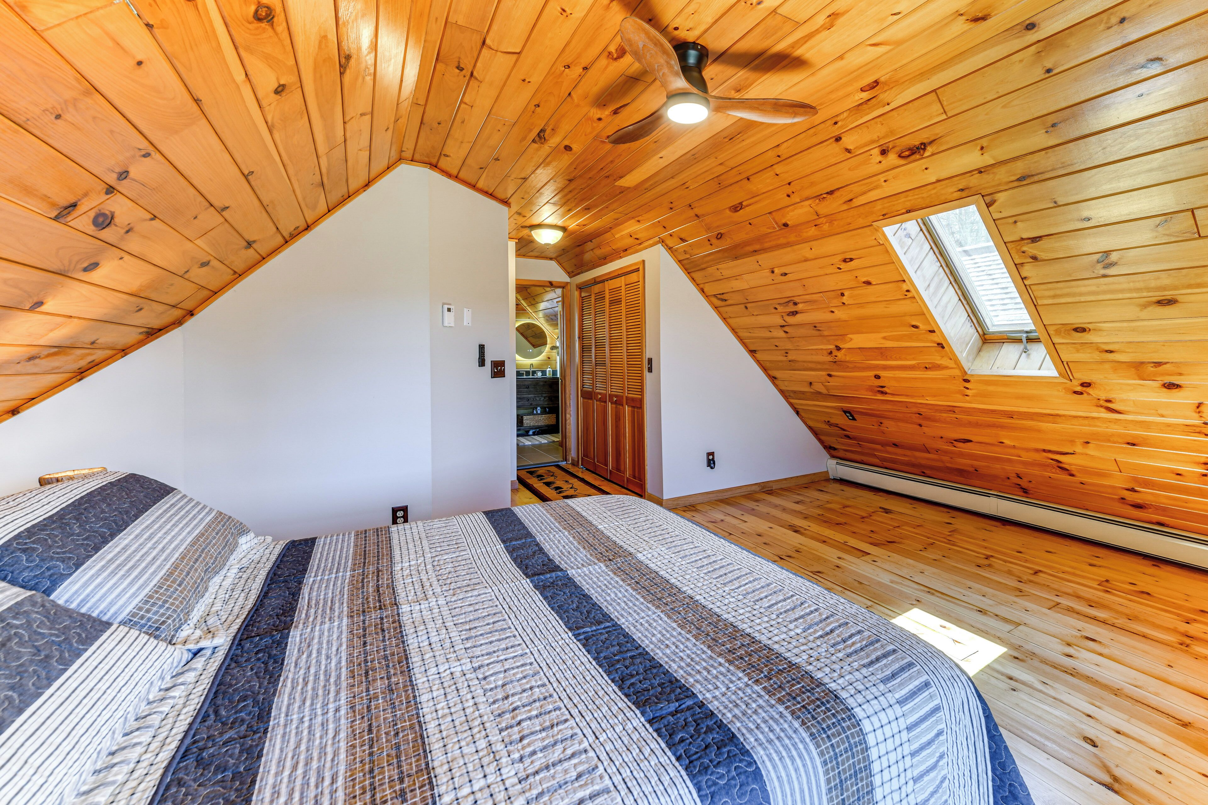 Un dormitorio con techo inclinado, paredes de madera, cama con ropa de cama estampada, ventilador de techo y tragaluz.