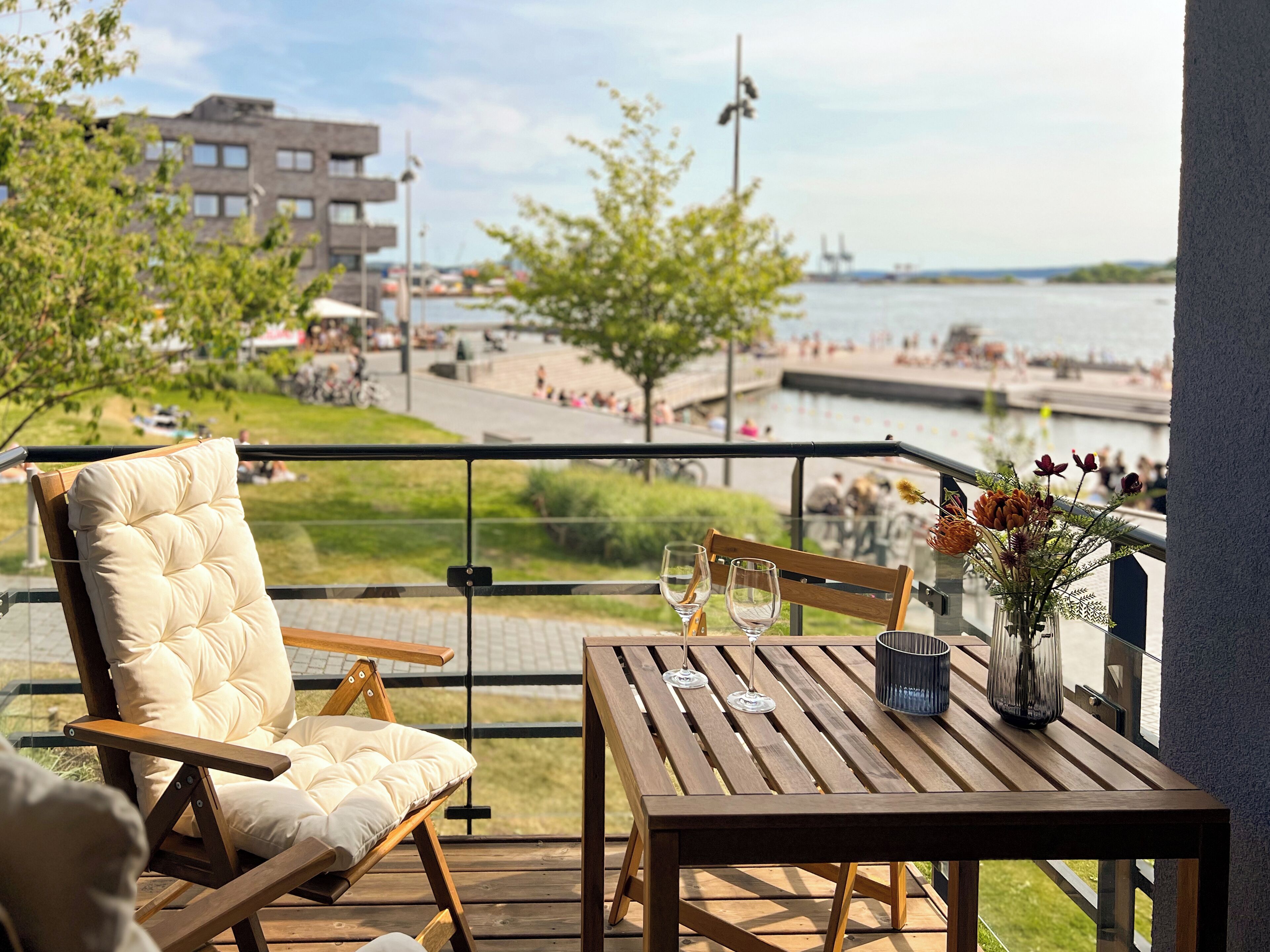 A balcony with a cushioned chair and a wooden table, overlooking a riverside promenade with people and buildings in the background.